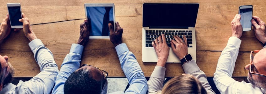 four business people sit at desk with different electronic devices